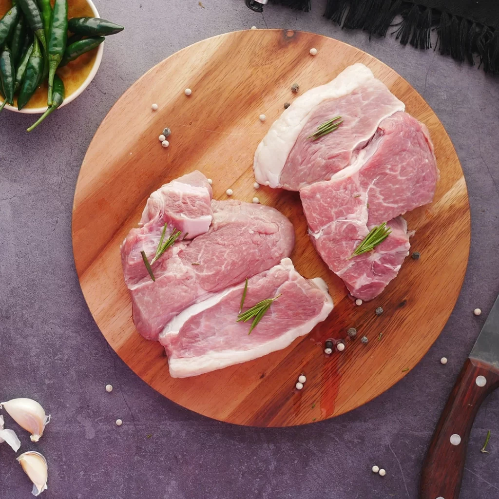 Three raw pork steaks on a round wooden board, garnished with rosemary sprigs, surrounded by garlic cloves, green peppers, and a chef's knife on a gray countertop.