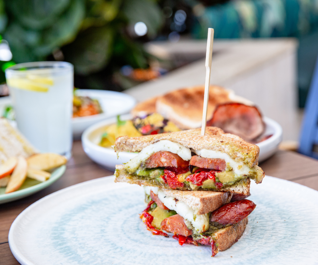 A stacked sandwich with sausage, mozzarella, pesto, and roasted tomatoes is served on a white plate with a skewer. Other dishes and a glass of lemon water are in the background.