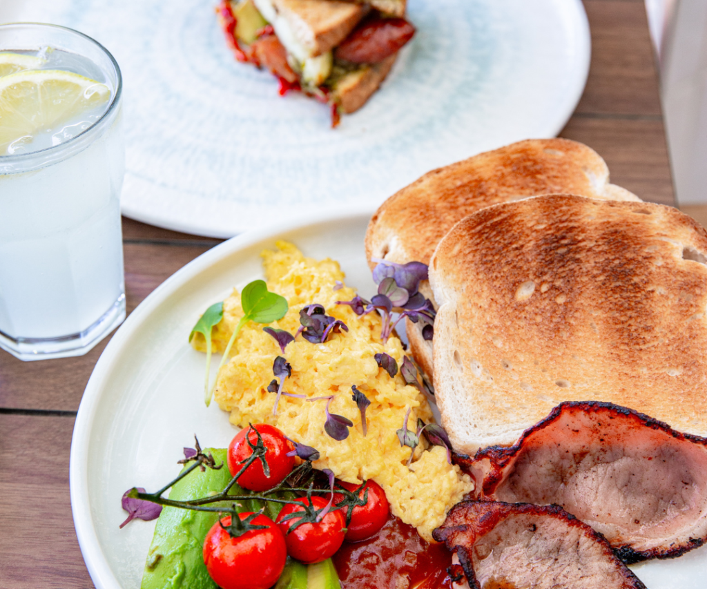 A breakfast plate with scrambled eggs, toast, grilled tomatoes, avocado, bacon, and microgreens. A glass of lemon water and a sandwich are in the background.