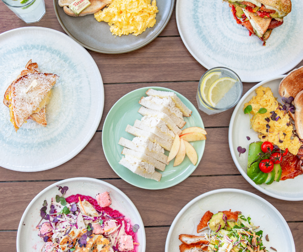 A top view of various breakfast dishes, including sliced bread, sandwiches, scrambled eggs, fruit, and colorful salads, arranged on a wooden table.