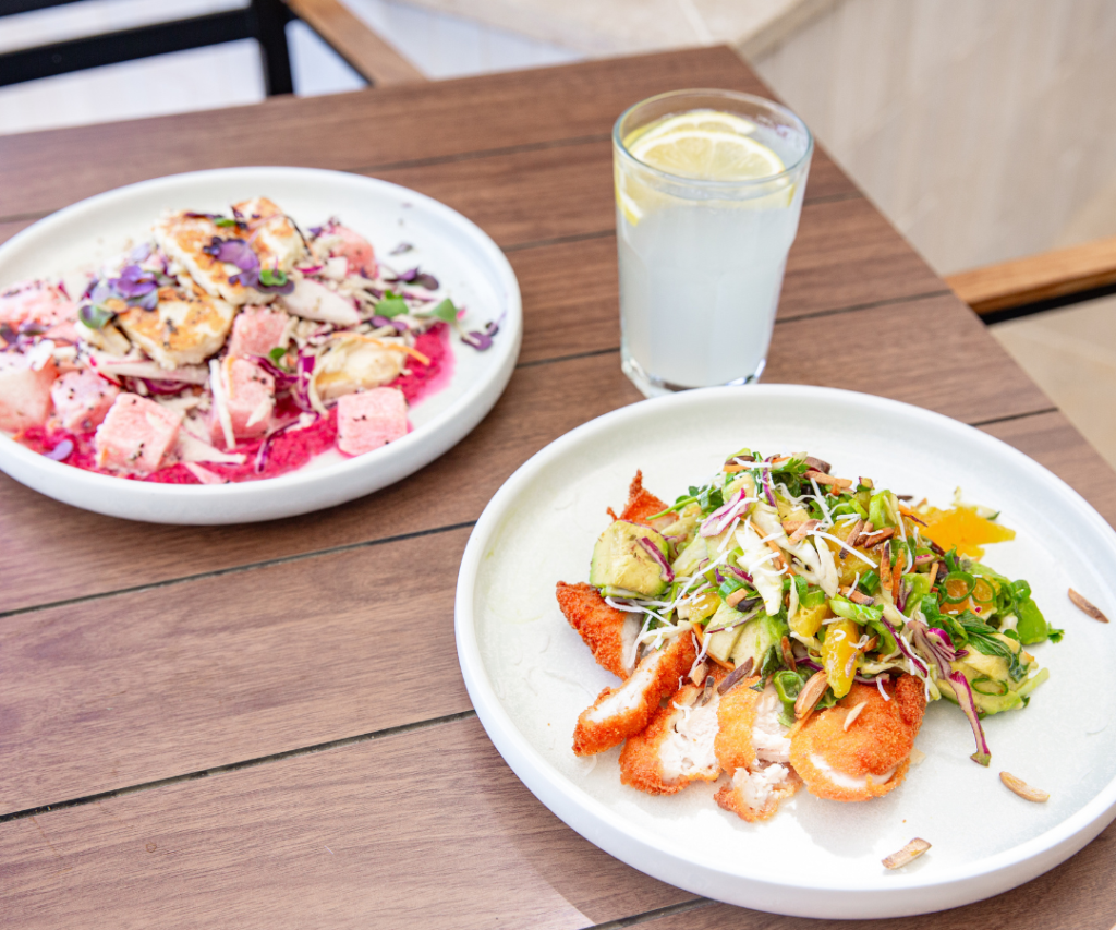 Two plates of salad with grilled chicken and vegetables on a wooden table, next to a glass of lemonade with a slice of lemon.