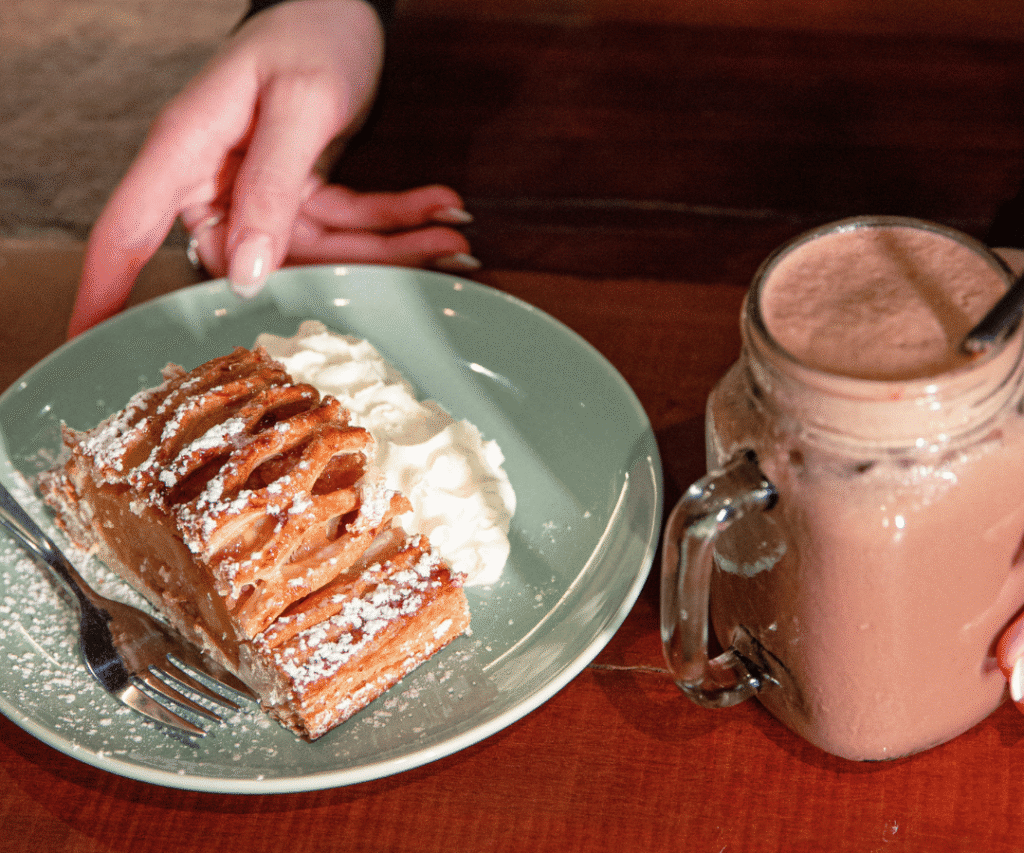A person’s hands are near a plate with apple pie and whipped cream, and a glass jar filled with chocolate milkshake on a wooden table.