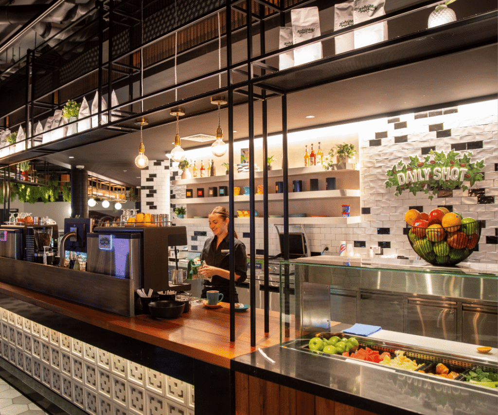 A barista prepares a drink behind a modern café counter with fruit bowls, glass display, hanging lights, and “Daily Shot” sign on a tiled wall.