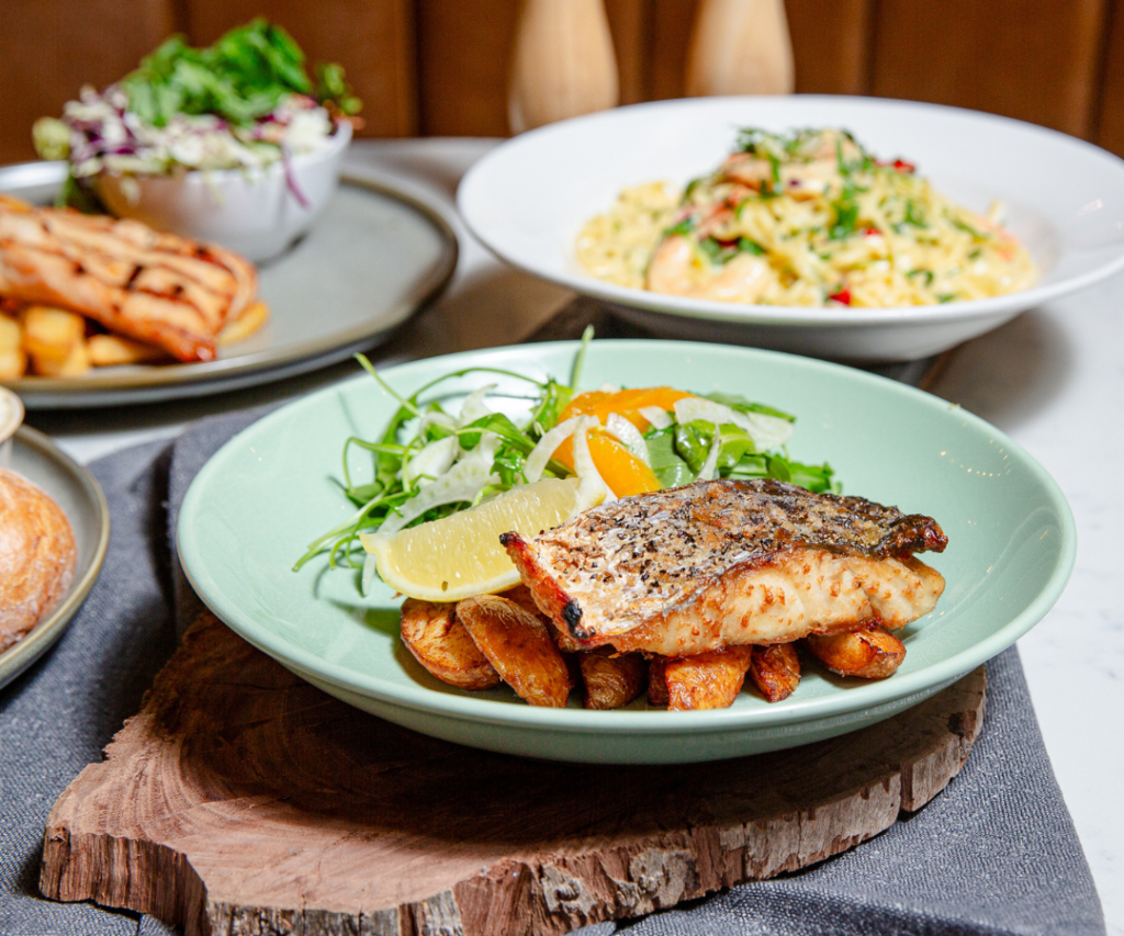 A plate with grilled fish fillet on potato wedges, garnished with lemon slices and greens; pasta and salad in bowls are in the background.