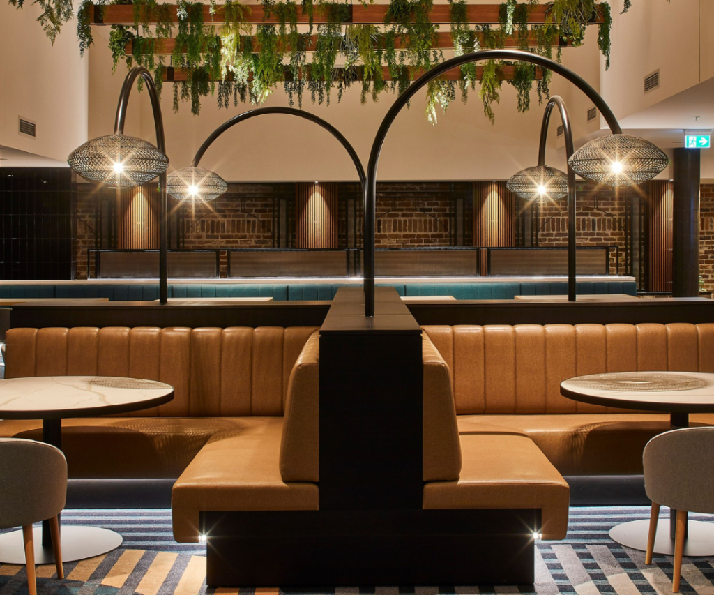 Modern restaurant interior with tan upholstered booth seating, round tables, overhead pendant lights, greenery hanging from the ceiling, and exposed brick wall in the background.