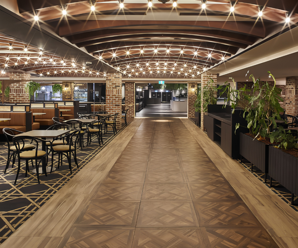 A modern, well-lit restaurant interior with wooden floors, geometric patterns, black chairs, potted plants, and exposed brick walls under a ceiling with decorative lighting.