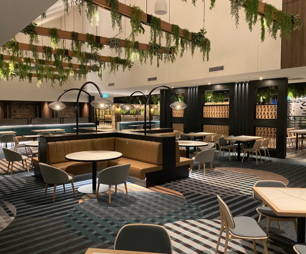 Modern restaurant interior with empty tables and chairs, hanging plants from the ceiling, and patterned carpet flooring. Soft lighting creates a calm atmosphere.