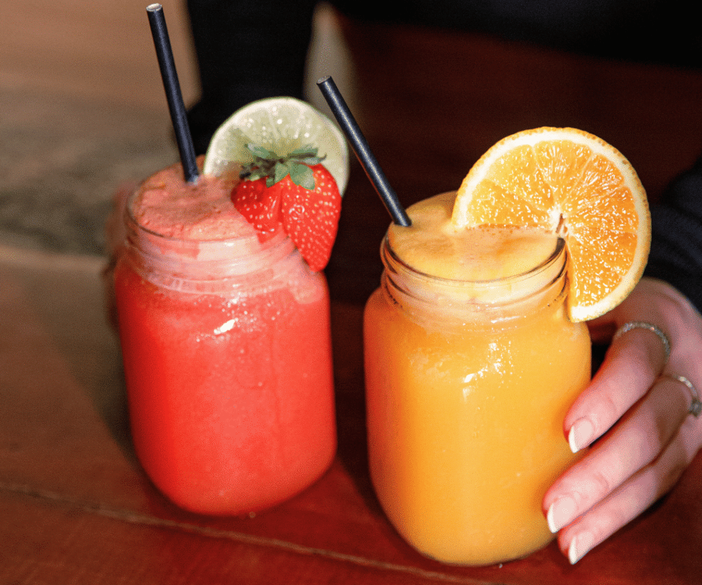 Two mason jars with colorful smoothies, one red with a strawberry and lime garnish, and one orange with an orange slice garnish, each with a straw on a wooden table.
