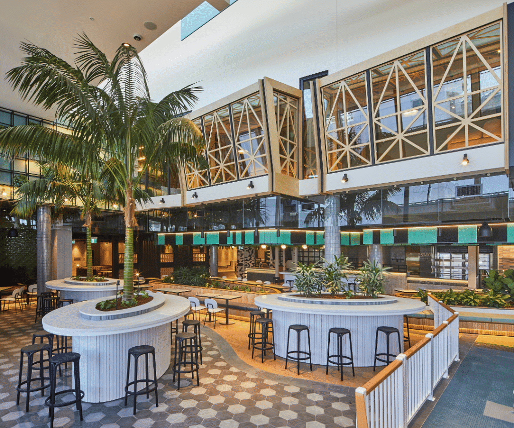 Modern indoor dining area with round tables, black stools, potted plants, palm tree, hexagonal tile floor, and enclosed upper balcony seating. Skylight and large windows provide natural light.