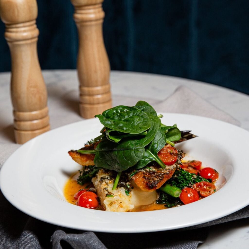 Grilled fish fillet topped with fresh spinach, served with cherry tomatoes, broccolini, and a light sauce on a white plate. Two wooden pepper mills in the background.