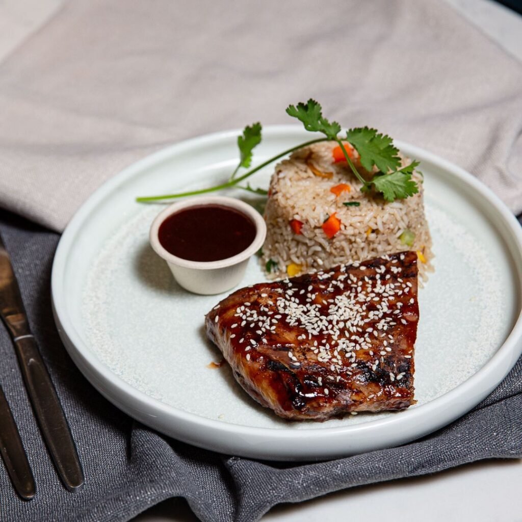 A white plate with glazed meat topped with sesame seeds, a serving of fried rice with vegetables, a small cup of sauce, and a sprig of cilantro for garnish.