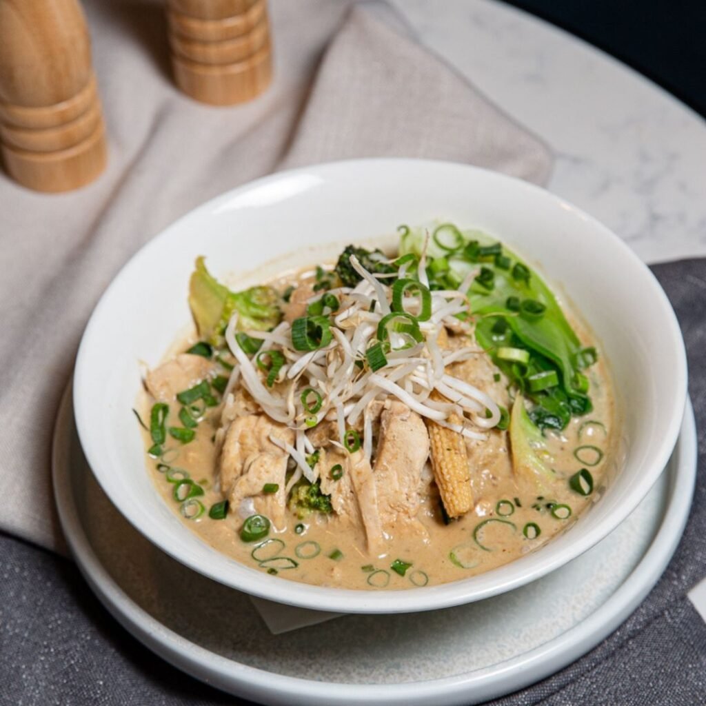 A bowl of ramen with sliced chicken, baby corn, bok choy, bean sprouts, and chopped green onions in a creamy broth, served on a white plate.