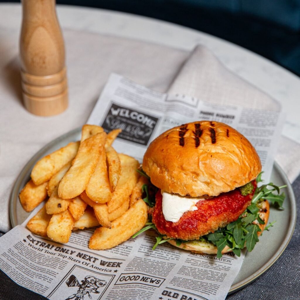 A breaded chicken burger with lettuce and sauce, served with thick-cut fries on a plate lined with newspaper print, on a table with a pepper mill in the background.