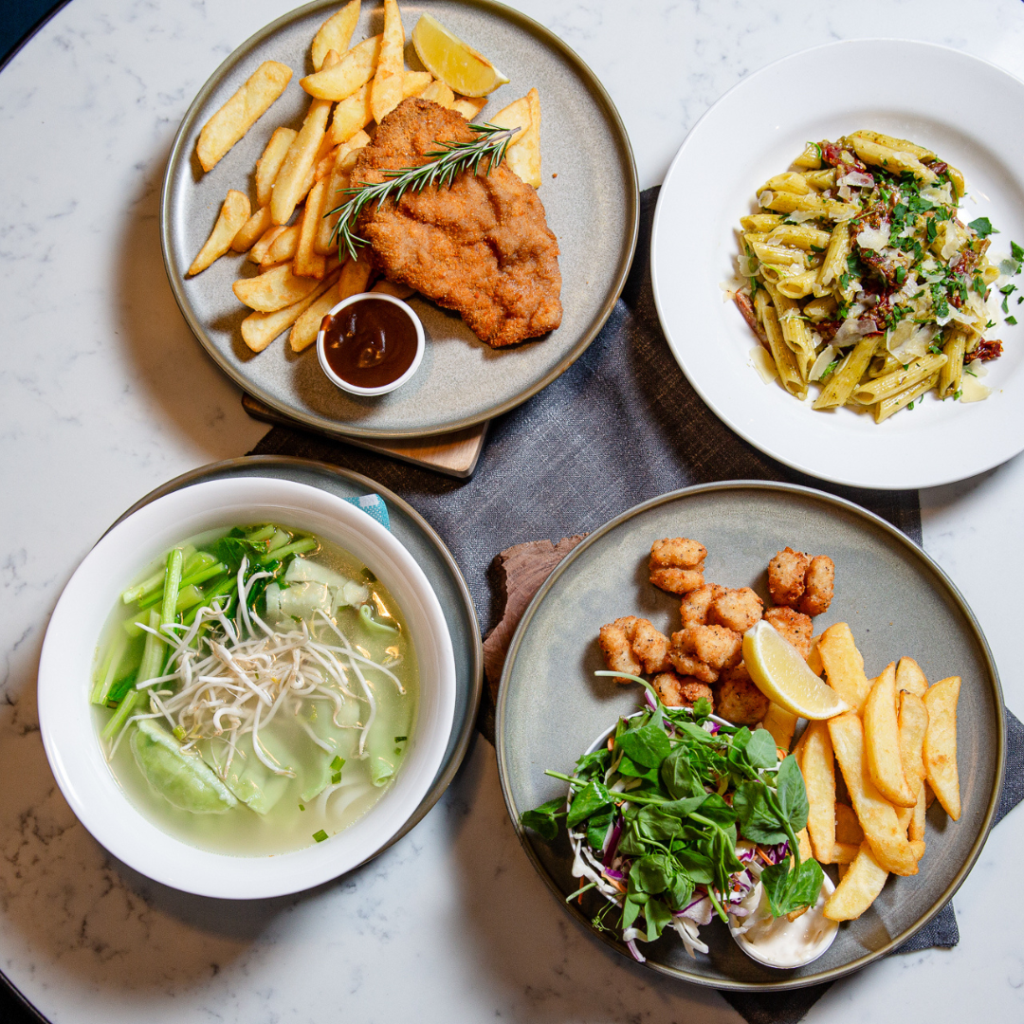 Four plates of food on a round white table: fried fish with fries, pasta with greens, noodle soup with vegetables, and fried bites with salad, fries, and lemon wedges.