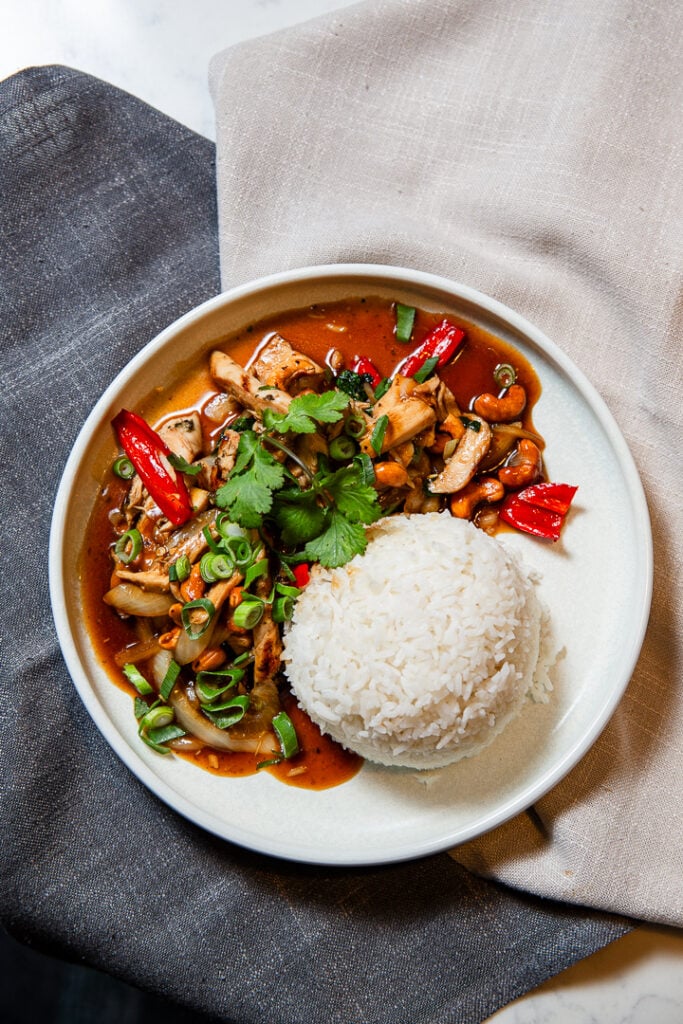 A plate with a serving of white rice and stir-fried vegetables with tofu in a brown sauce, garnished with cilantro, green onions, and red chili peppers.