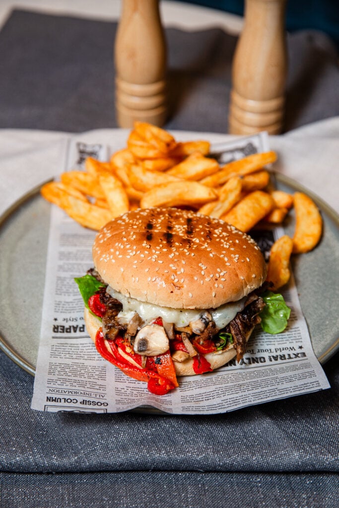 A sesame seed burger with lettuce, mushrooms, and red peppers is served on a plate with crinkle-cut fries over a sheet of printed paper.