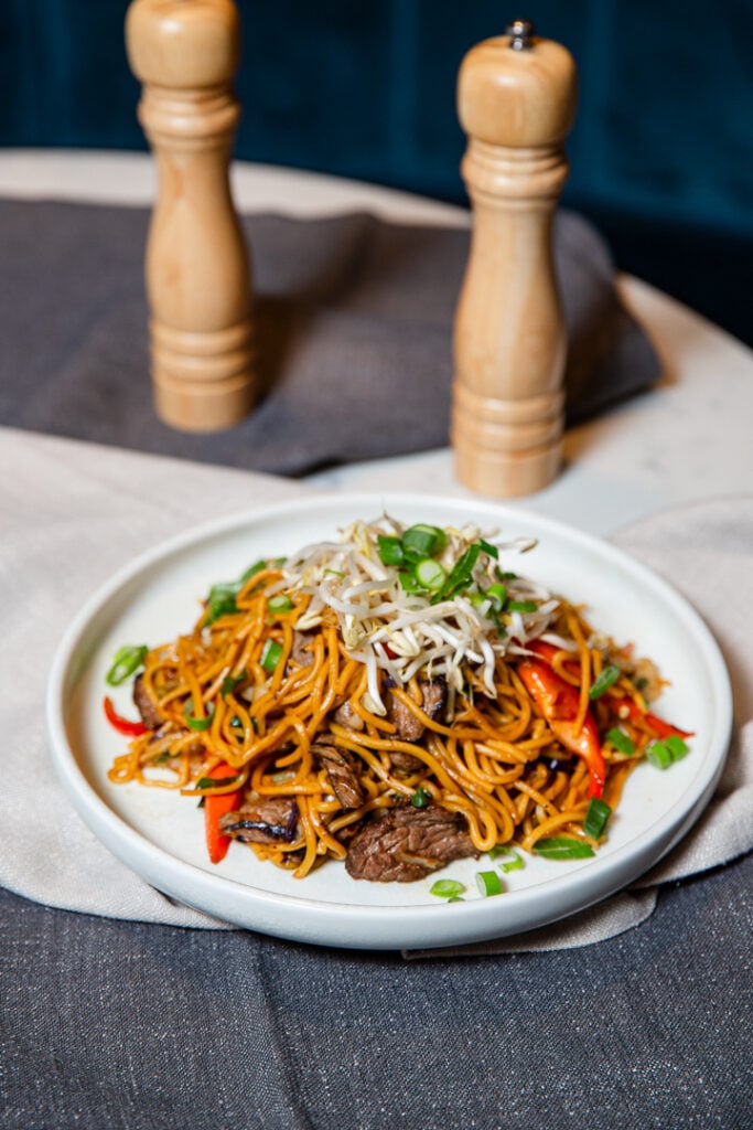 A plate of stir-fried noodles with beef, bell peppers, scallions, and bean sprouts, placed in front of two wooden pepper mills on a table.