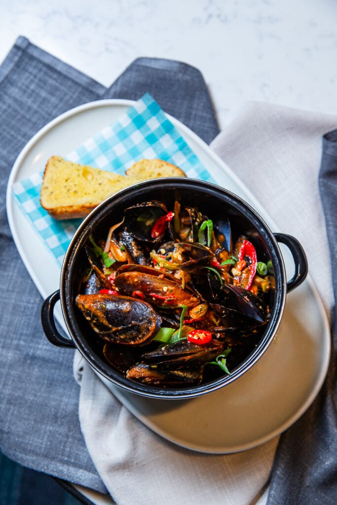 A black bowl of cooked mussels with vegetables and herbs, served on a plate with two slices of bread, on a fabric napkin.