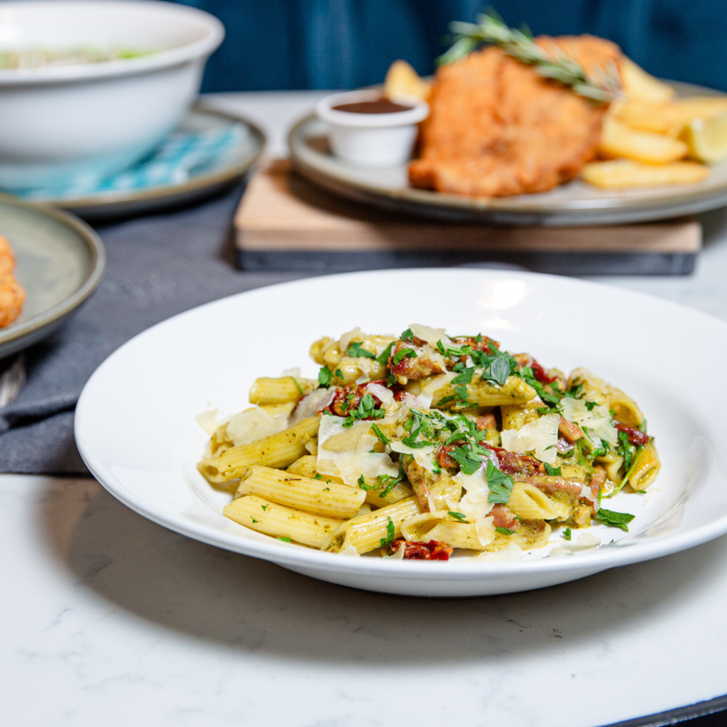 A white plate with penne pasta in a creamy sauce, topped with herbs, artichokes, and sun-dried tomatoes, sits on a marble table alongside other dishes in the background.