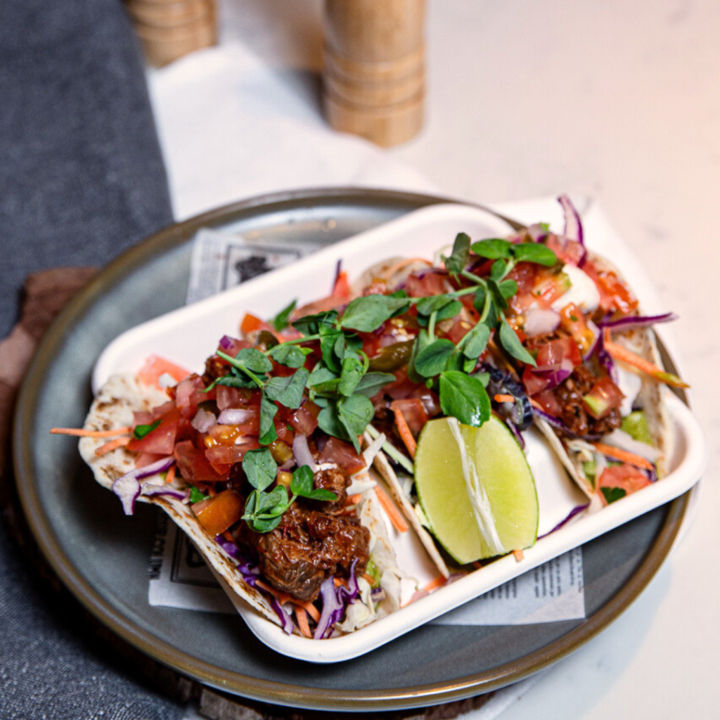 Two soft shell tacos filled with shredded meat, diced tomatoes, slaw, greens, and a lime wedge, served on a small tray atop a round plate.