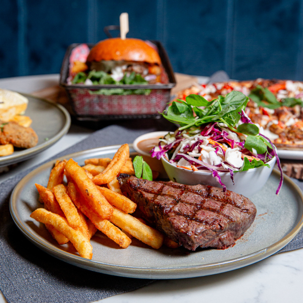A plate with grilled steak, French fries, and a side salad in the foreground; other dishes including a burger and pizza are in the background.