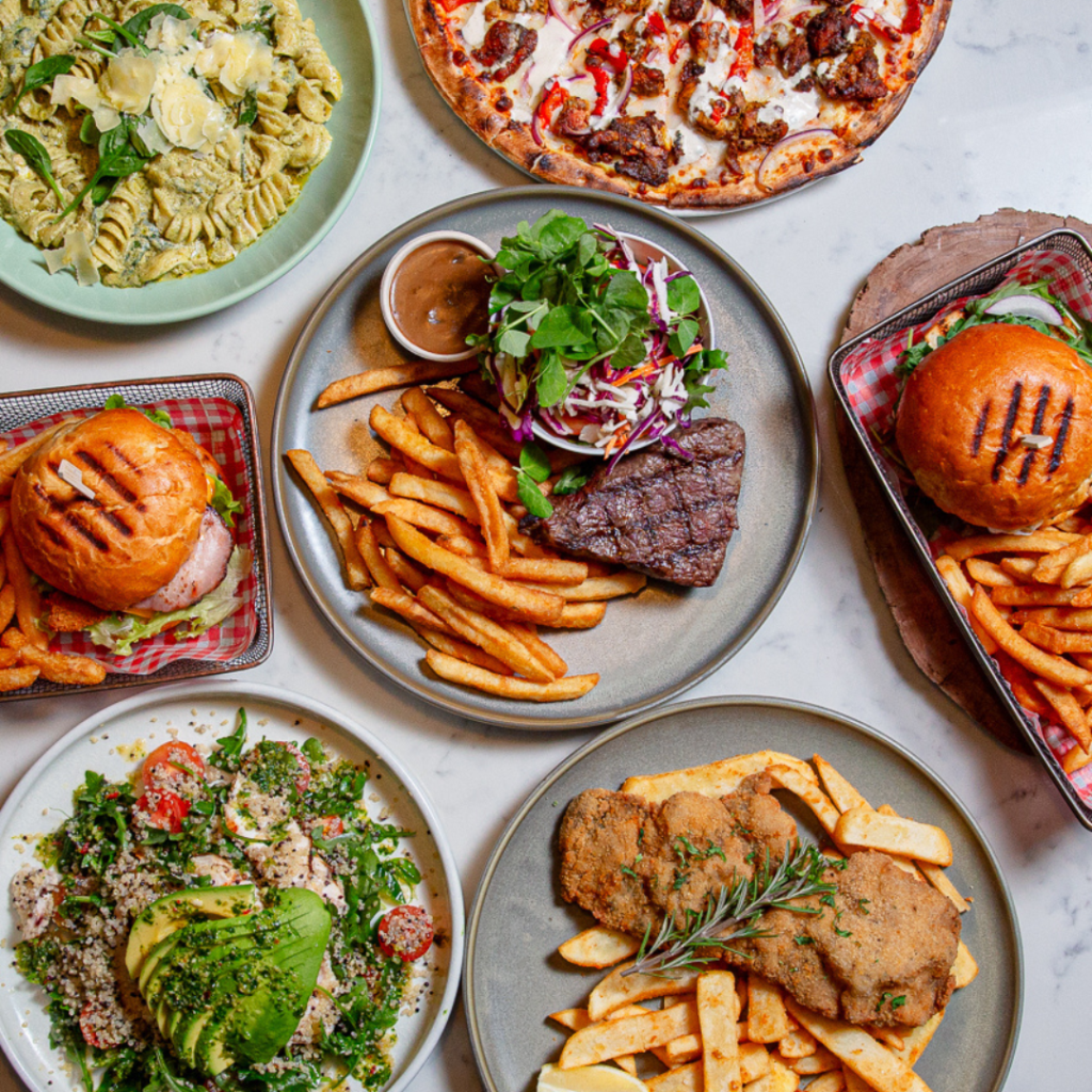 A table with various dishes including burgers with fries, a steak with fries and salad, schnitzel with fries, pasta, pizza, and a salad with avocado.