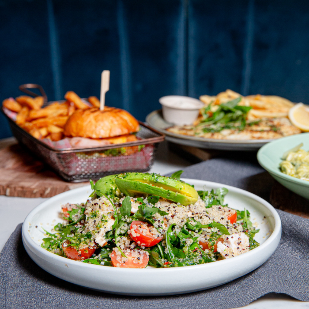 A plate of salad with quinoa, cherry tomatoes, arugula, and avocado sits in the foreground, with a burger and fries and other dishes blurred in the background.