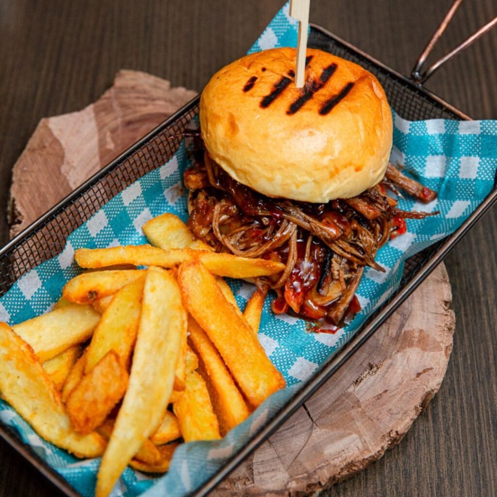 A pulled pork sandwich with barbecue sauce and a side of thick-cut fries served in a metal basket with blue checkered paper.