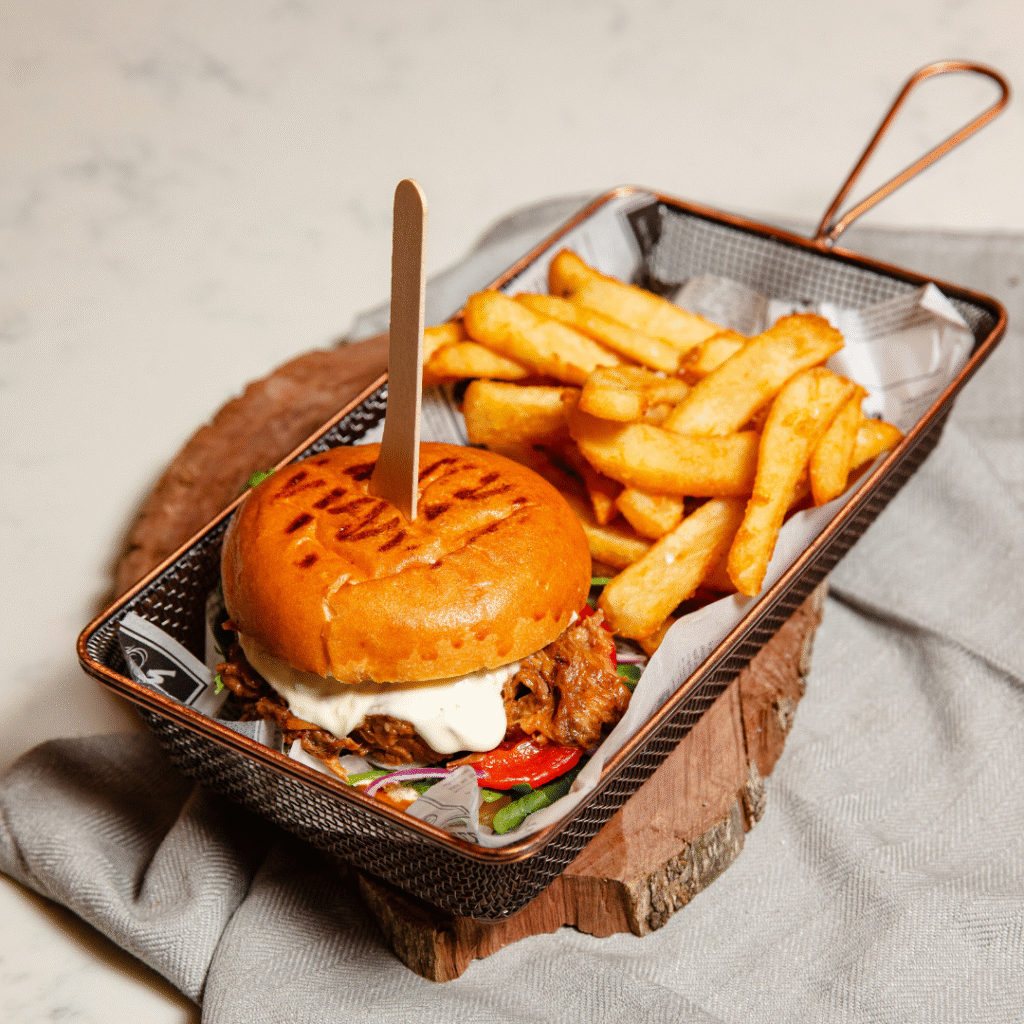 A burger with lettuce, sauce, and a bun marked “VEGAN” is served with thick-cut fries in a small basket on a wooden slab.