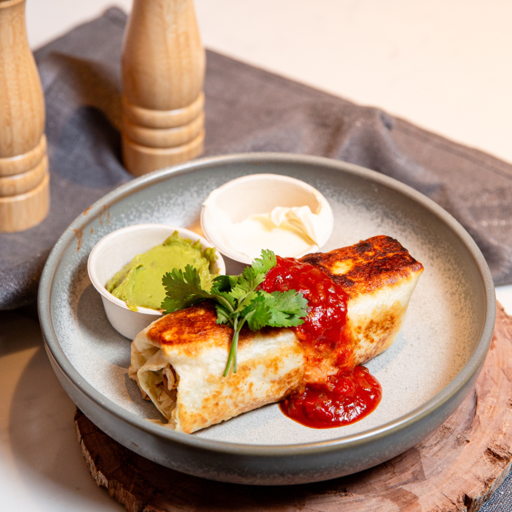 A grilled burrito topped with salsa and cilantro, served with sides of guacamole and sour cream on a gray plate. Two wooden pepper mills are in the background.