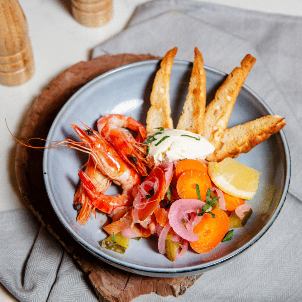 A plate with cooked shrimp, cherry tomatoes, pickled onions, a dollop of cream, lemon wedge, and three slices of toasted bread on a gray napkin.
