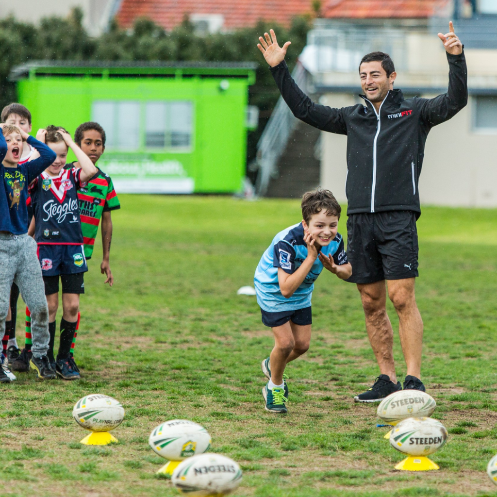 Children participate in a rugby training session outdoors with a coach. One child runs between rugby balls while others watch and react. The coach signals with raised arms.