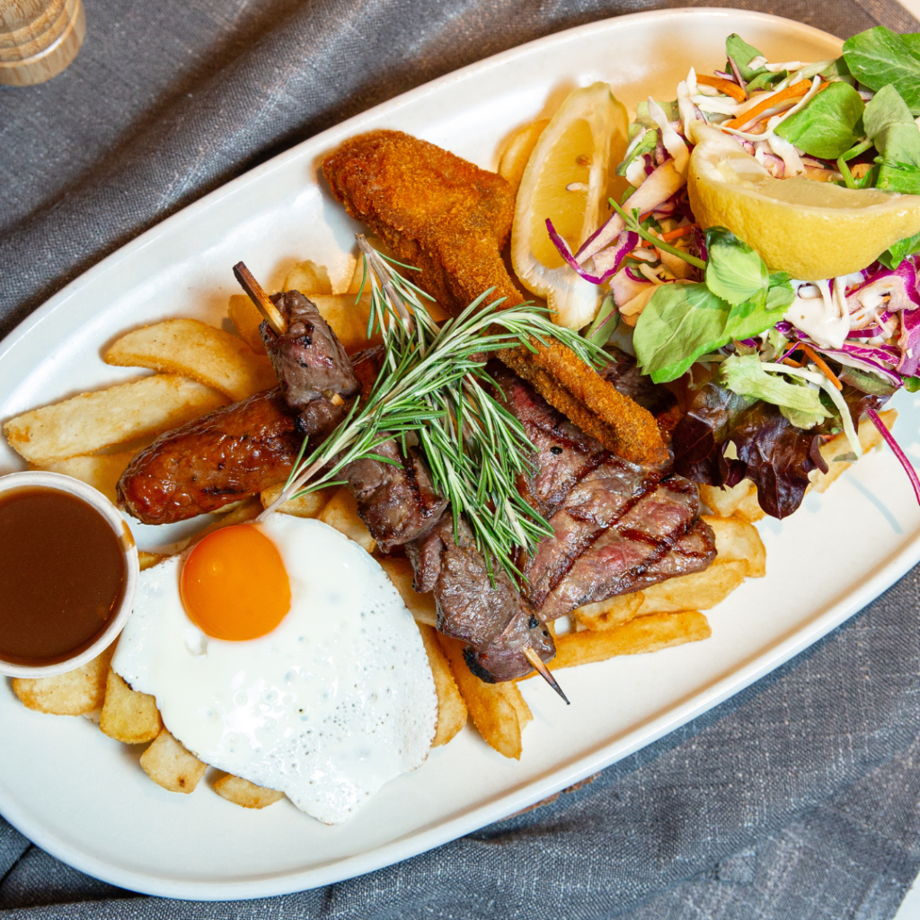 A plate with steak skewers, sausage, a fried egg, fries, brown sauce, crumbed fish, and a side salad with lemon wedge on a white oval dish.