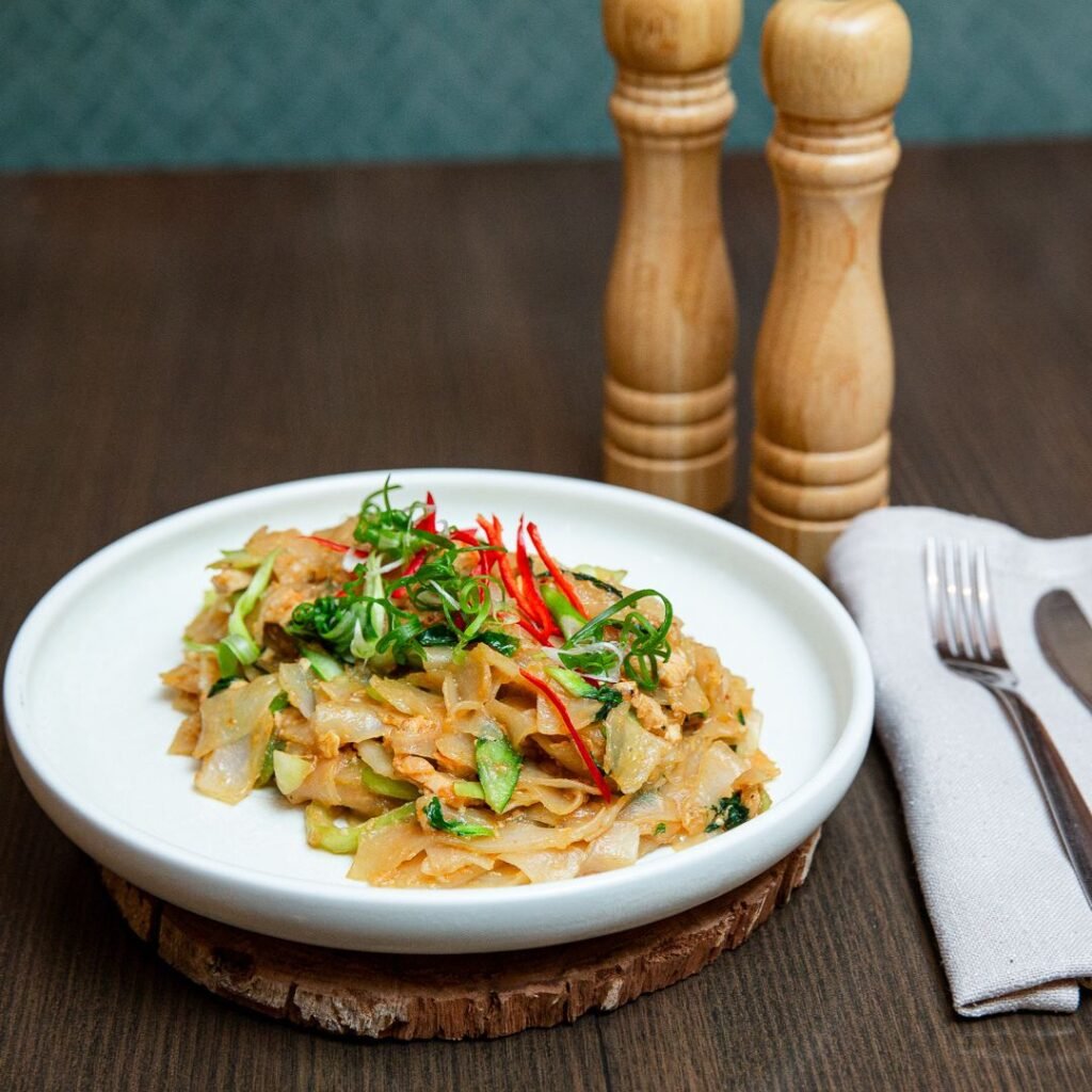 A plate of stir-fried noodles with vegetables and garnished with sliced chilies and herbs, placed on a wooden table beside salt and pepper grinders, fork, and knife.