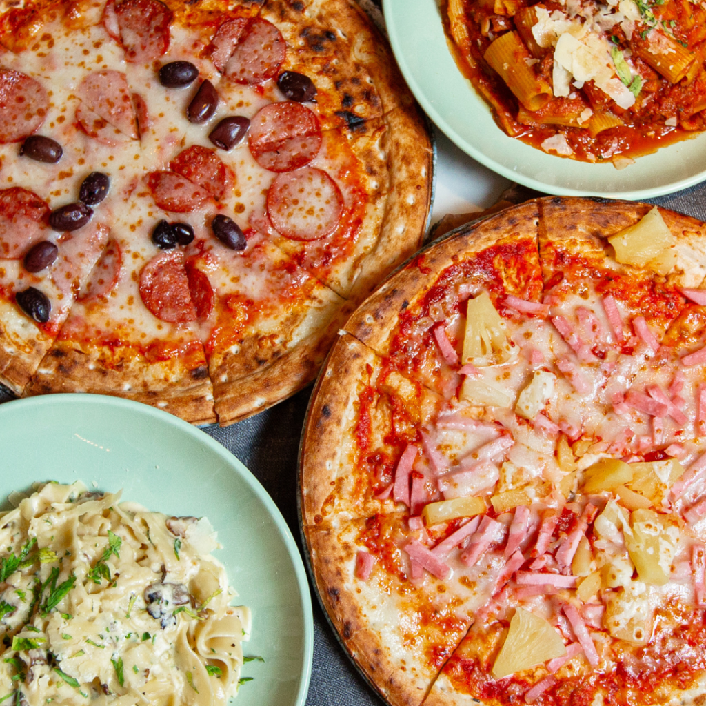 Three pizzas with different toppings and a plate of creamy pasta are arranged on a table with green plates.
