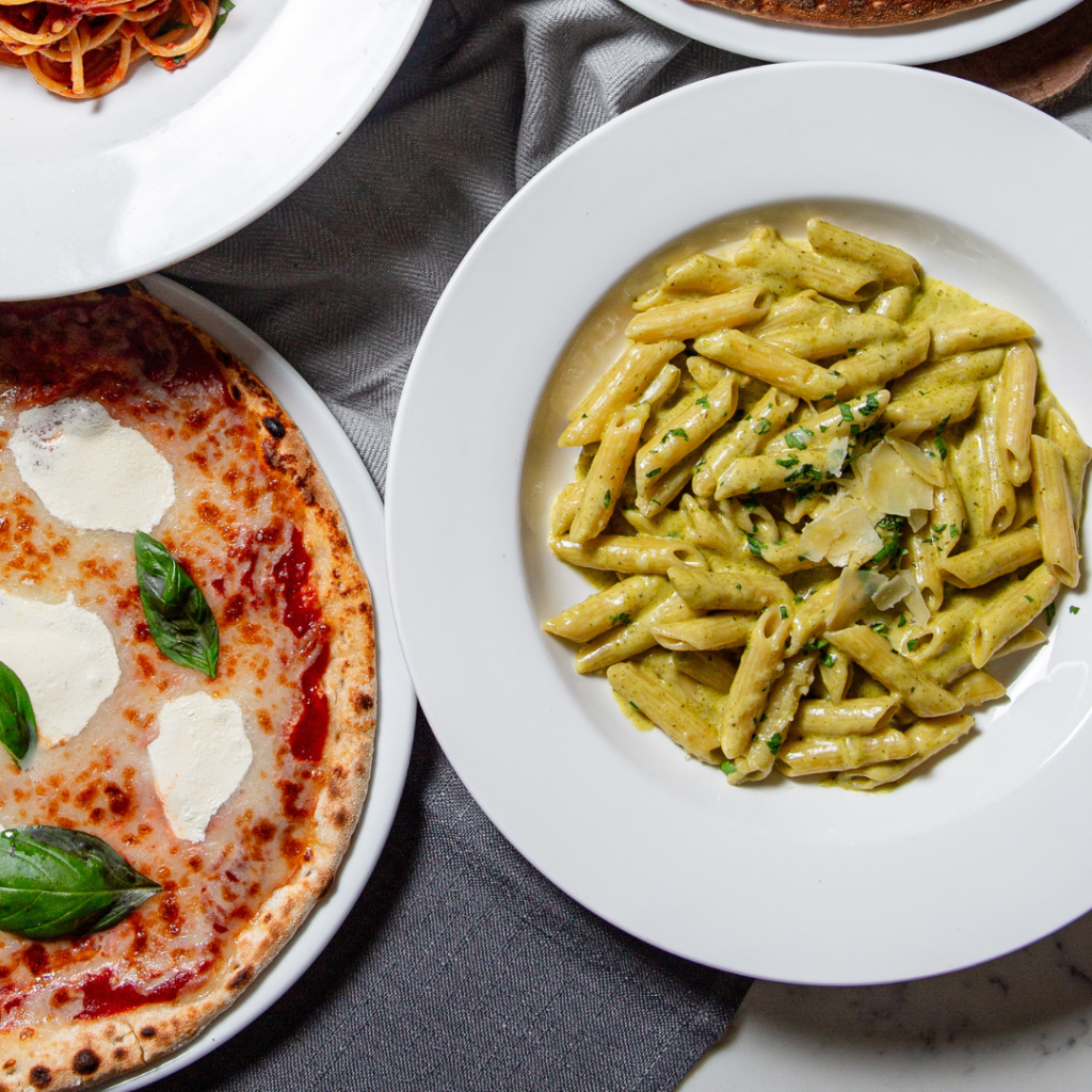 A white plate of penne pasta with pesto sauce and shaved parmesan, next to a margherita pizza and a plate of spaghetti.