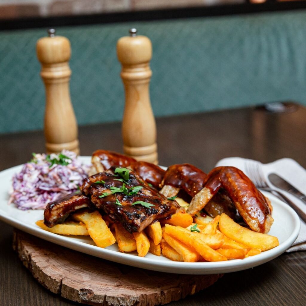 A plate of barbecued ribs, sausage, thick-cut fries, and coleslaw is served on a wooden board, with salt and pepper grinders in the background.