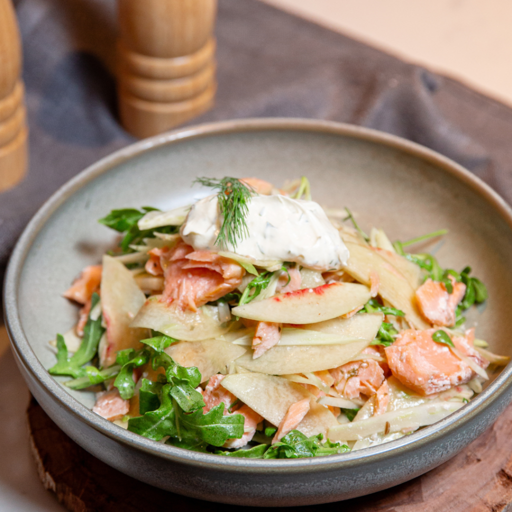 A salad with arugula, apple slices, and smoked salmon topped with a dollop of creamy dressing and a sprig of dill, served in a gray bowl.