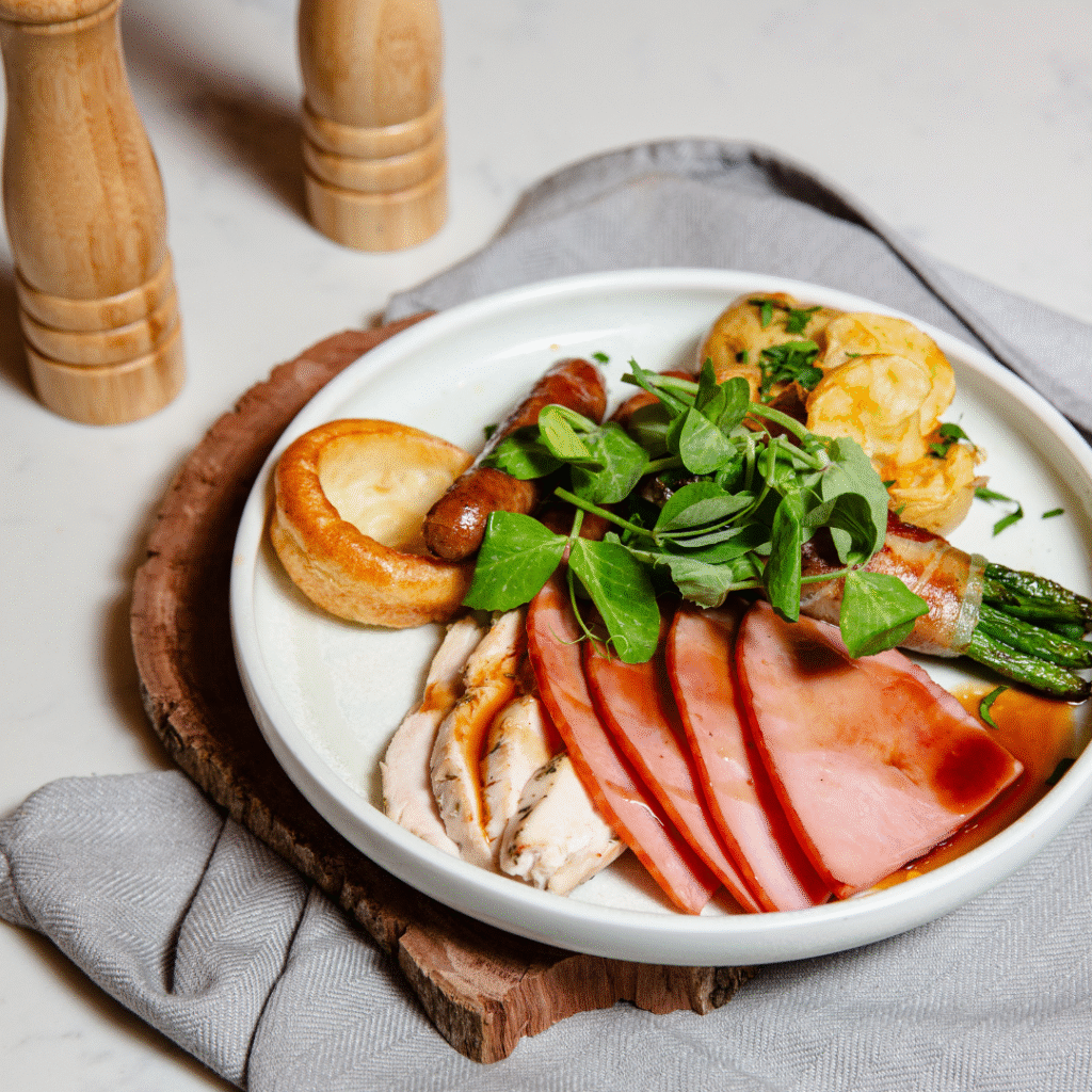A plate with sliced ham, roast turkey, sausages, green vegetables, Yorkshire pudding, and a garnish, placed on a wooden board with a napkin.