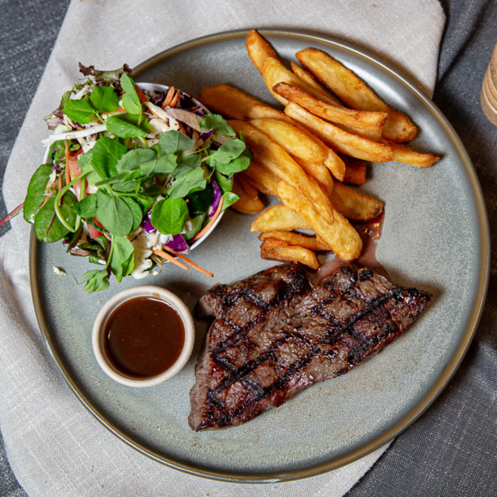 A plate with grilled steak, thick-cut fries, a small bowl of fresh mixed salad, and a cup of brown sauce, served on a light gray plate.