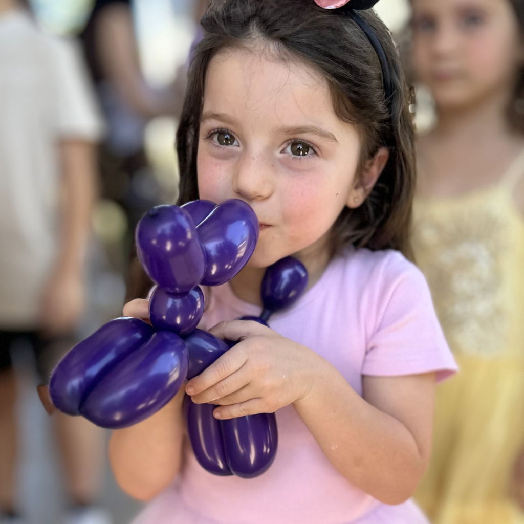 Young girl in a pink shirt holds and kisses a purple balloon animal, with other children visible in the blurred background.