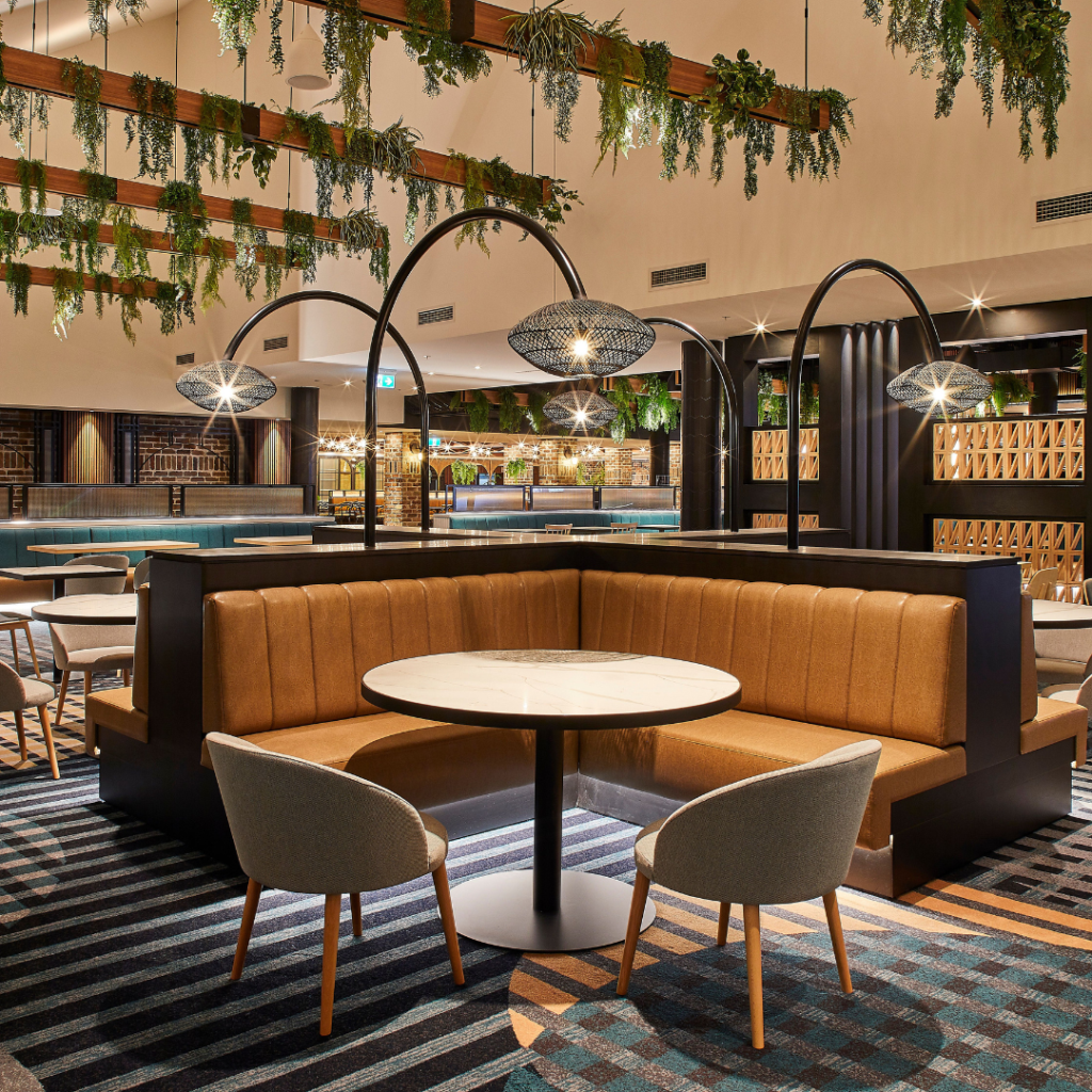 Modern restaurant interior with a round marble table, tan upholstered booth seating, gray chairs, patterned carpet, and hanging greenery with pendant lights.