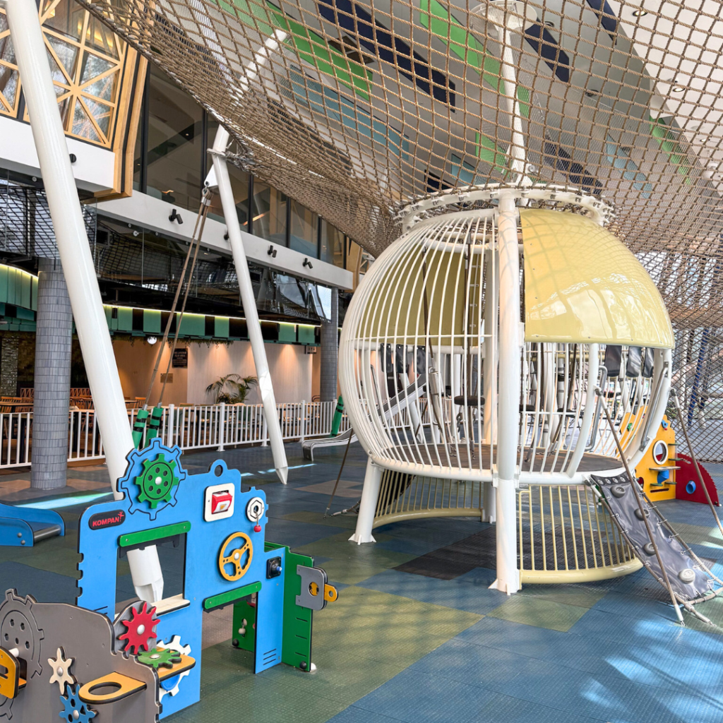 Indoor playground with climbing nets, a spherical play structure, a slide, and interactive panels, set on a cushioned floor in a spacious, well-lit area.