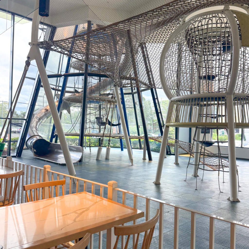 Indoor play area with climbing nets, a slide, and large play structures next to a seating area with wooden tables and chairs, enclosed by a white fence.