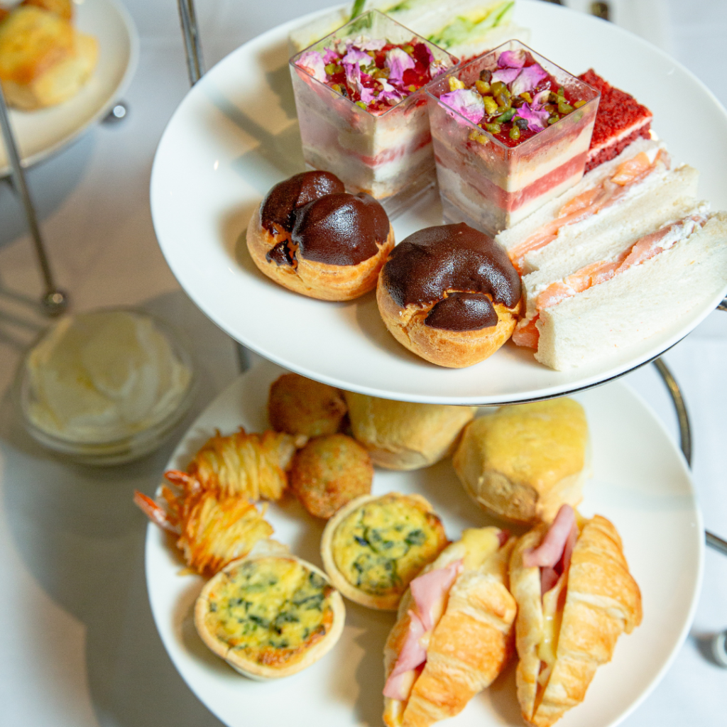 A two-tiered tray with assorted pastries, finger sandwiches, mini quiches, croissants, and layered desserts arranged for afternoon tea.