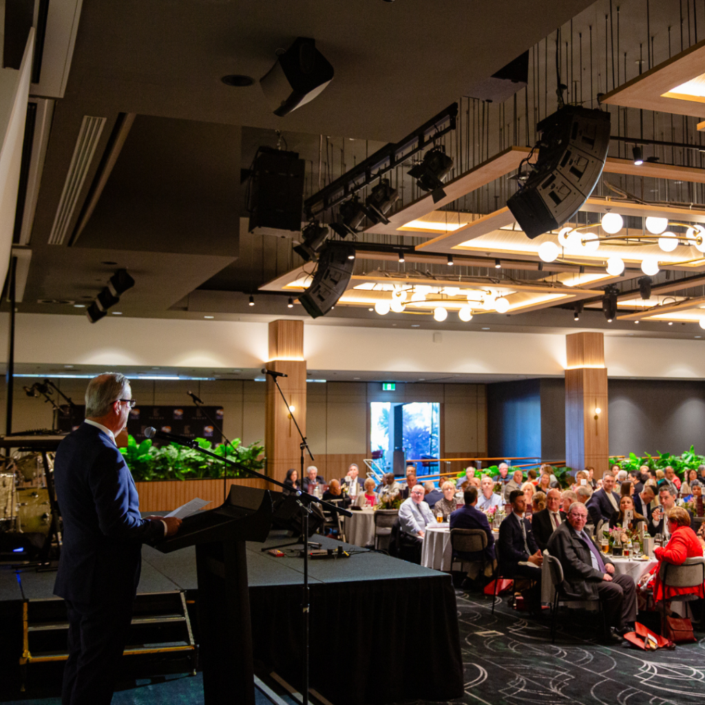 A speaker addresses an audience seated at round tables in a modern, well-lit conference room with decorative lighting and plants.