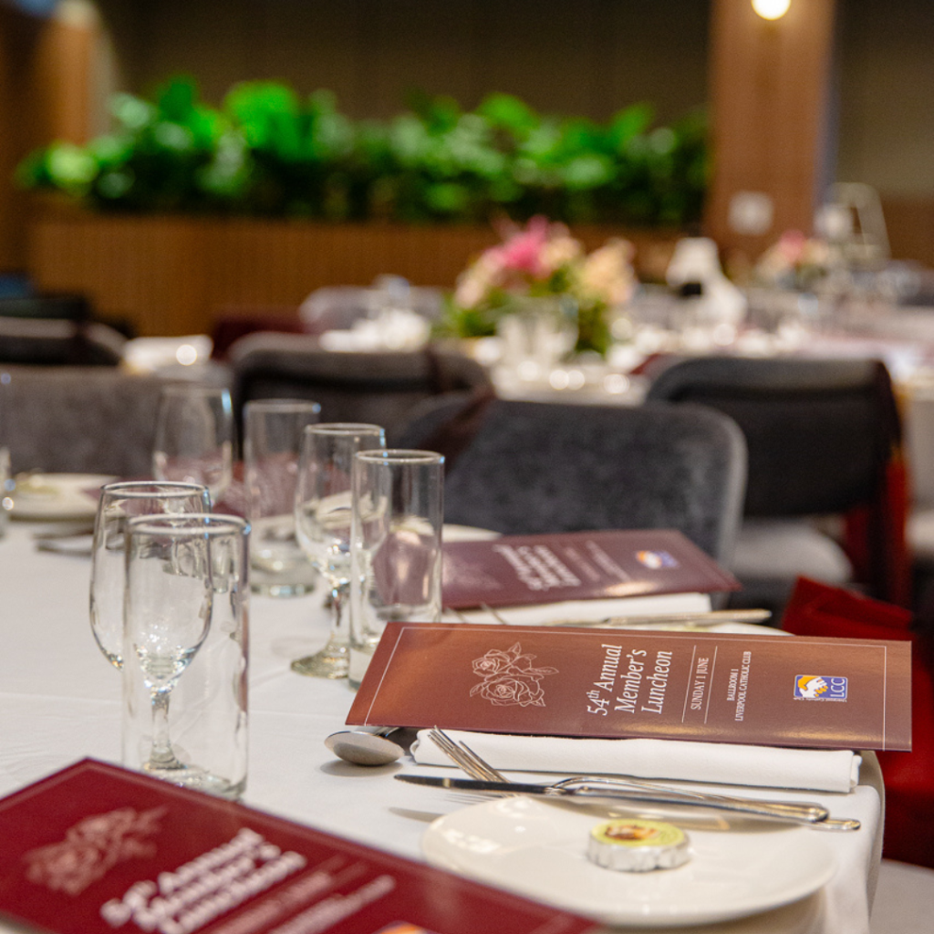 Table set for an event with glassware, cutlery, and maroon event programs titled "52nd Annual Member Luncheon"; green plants and floral arrangements in background.