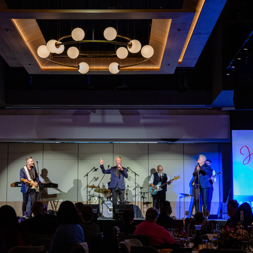 A band performs live on stage under modern lighting, with an audience seated at tables in the foreground.