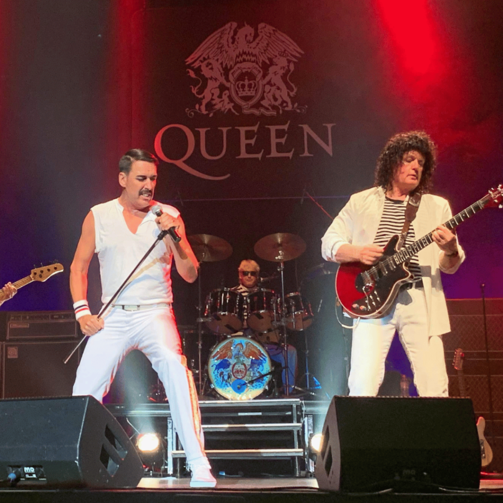 A band performs on stage with a "Queen" logo in the background; the singer holds a microphone, the guitarist plays, and a drummer is visible behind them. Red and purple stage lights shine.