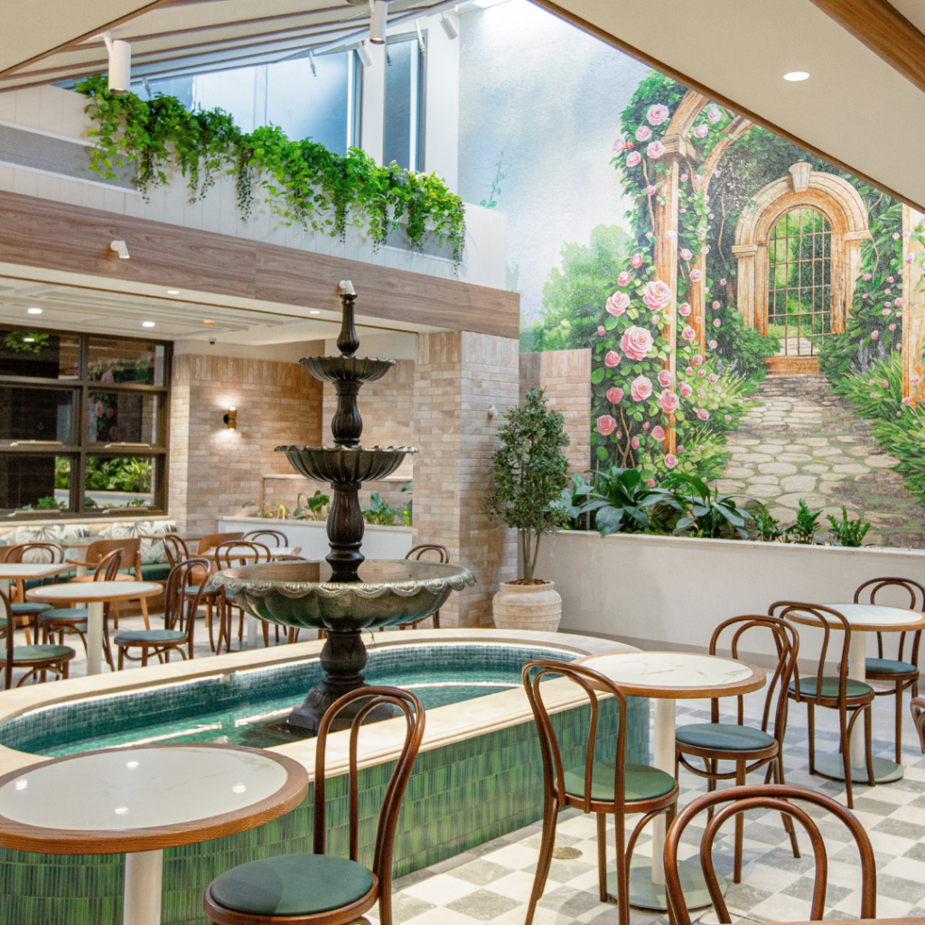 Indoor café with round tables, wooden chairs, a central tiered fountain, leafy plants, and a mural depicting a garden archway path on the far wall.